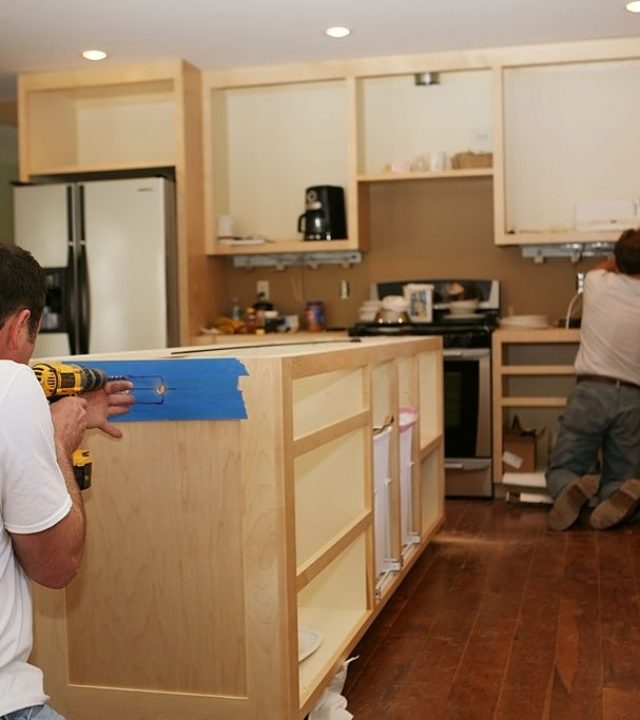 Cabinet maker installing custom made maple cabinets to kitchen island, while electrician installs under-cabinet lighting. 

The cabinet maker is cutting holes for electrical outlets and switches.

Very shallow DOF quickly drops off to nice soft focus.